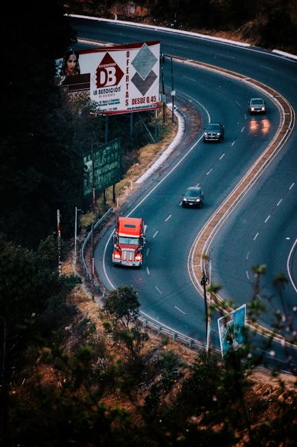 Aerial view of a winding dual carriageway highway with four visible vehicles, including a red truck in the foreground and three smaller cars, driving along the road surrounded by greenery and trees. Large roadside billboards are present, one advertising a local business with a woman's portrait, and another with promotional content. The scene is illuminated by natural daylight, highlighting the road's curves and the vehicles' movement. This image relates to route planning and transportation important for home relocation and furniture transport services, as provided by Man with Van Upton, emphasizing efficient moving routes to avoid delays during house removals.