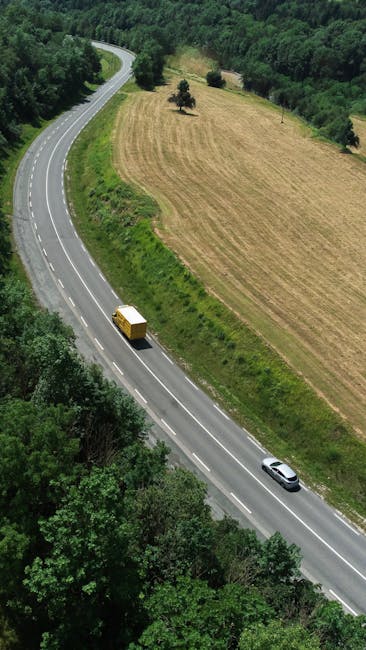 An aerial view of a winding two-lane road cutting through a rural landscape with lush green trees and open fields. A yellow moving box is placed on the rear of a small van traveling along the road, suggesting a home relocation or furniture transport in progress. The scene is illuminated by daylight, with the road bordered by clusters of trees on one side and cultivated fields on the other. This image highlights the importance of choosing optimal routes for efficient house removals, as supported by Man with Van Upton, particularly for destinations like Upton around CH49, where careful planning of moving logistics helps avoid delays during packing and loading processes.