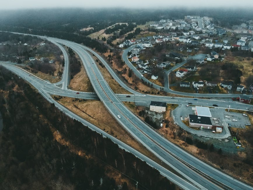 Aerial view of a winding dual carriageway highway with four visible vehicles, including a red truck in the foreground and three smaller cars, driving along the road surrounded by greenery and trees. Large roadside billboards are present, one advertising a local business with a woman's portrait, and another with promotional content. The scene is illuminated by natural daylight, highlighting the road's curves and the vehicles' movement. This image relates to route planning and transportation important for home relocation and furniture transport services, as provided by Man with Van Upton, emphasizing efficient moving routes to avoid delays during house removals.