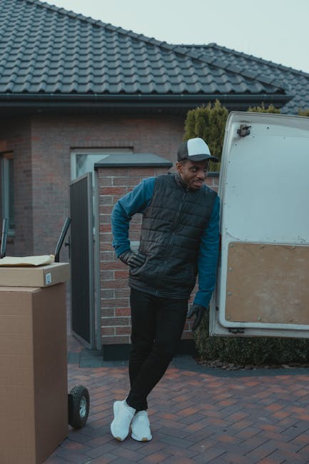 A man dressed in a blue long-sleeve top, black vest, and black pants, wearing a white cap and gloves, stands next to the open rear door of a white van during a home relocation. He appears to be waitin