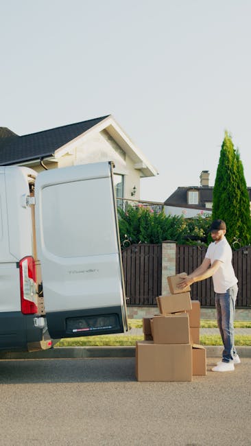 A man dressed in a blue long-sleeve top, black vest, and black pants, wearing a white cap and gloves, stands next to the open rear door of a white van during a home relocation. He appears to be waitin