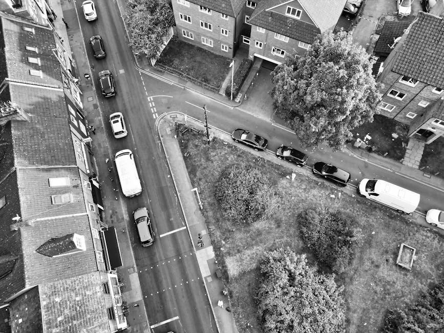 An aerial black-and-white photograph shows a narrow residential street with parked cars lining both sides, including a white van positioned at the entrance to a driveway. Adjacent to the street, there