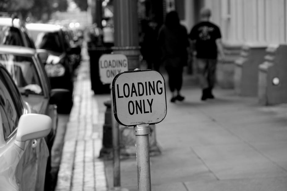 Black and white photograph of a street scene showing a row of parked cars along the curb with a focus on a metal sign reading 'Loading Only' positioned on a sidewalk post. Behind the sign, a van is partially visible with its rear doors open, indicating a loading process typical in house removals. In the background, three pedestrians walk past a building on the pavement, with some steps and architectural details visible on the buildings' facades. The scene captures an outdoor environment commonly associated with moving logistics, where proper loading zones and street loading permits, such as those possibly needed for a home relocation, are relevant. The lighting suggests daytime, and the overall composition emphasizes the importance of designated loading areas for efficient and legal furniture transport or packing and moving activities, as managed by professional removals services like Man with Van Upton.
