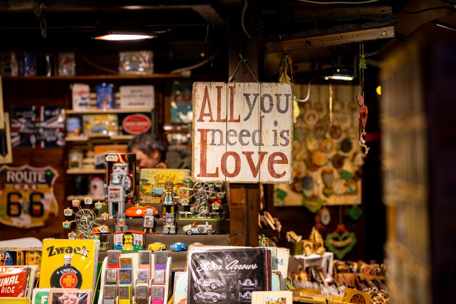 Inside a shop or storage area, there is a wooden sign hanging from the ceiling with the message 'All you need is Love' in large, colorful letters. Behind the sign, shelves stocked with vintage-style tin toys, figurines, and collectible items are visible, with some toys resembling robots and vehicles. Several small, decorative items and packaging materials such as cardboard boxes and plastic wrapping are arranged on the shelves. To the side, a man is partially visible, possibly involved in packing or sorting items as part of a house removal or relocation process. The environment features warm lighting and a cluttered yet organized appearance typical of a retail or storage space used for moving preparations. This setting is relevant to furniture transport, packing, and logistical aspects of home relocations, which Man with Van Upton handles professionally.