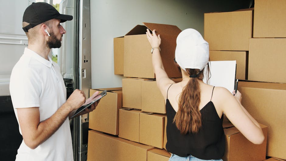 A man wearing a black cap, white t-shirt, and wireless earbuds stands beside an open moving van, holding a tablet and inspecting packed cardboard boxes. A woman with long reddish-brown hair tied back, wearing a white cap and black sleeveless top, is in the midst of loading or organising these boxes, some of which are open, revealing packing materials such as tissue paper and bubble wrap. The scene takes place on a driveway or loading area adjacent to a house, with additional cardboard boxes stacked nearby, ready for home relocation or furniture transport. The environment is well-lit with natural daylight, highlighting the process of packing and moving in a professional manner, with furniture blankets, hand trucks, or straps potentially visible as part of the loading process, supporting efficient moving logistics as managed by Man with Van Upton.