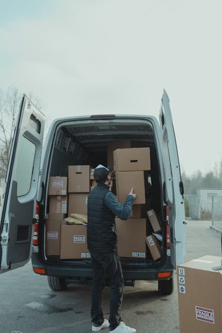 A person wearing a black vest, dark trousers, and a dark cap is standing next to the open rear doors of a large white moving van, which is parked outdoors on a paved surface. Inside the van, several cardboard boxes of various sizes are stacked, some sealed with packing tape and others partially visible, indicating a home relocation process. The boxes are arranged to optimize space within the van, ready for transportation. The individual is in the act of placing or adjusting a box, with their hand on one of the boxes near the opening. The environment appears to be an open area with a cloudy sky overhead, and a few trees or structures are visible in the distance. The scene is well-lit with natural light, emphasizing the practical nature of the furniture transport and packing involved in a professional removals service by Man with Van Upton, highlighting the loading process essential for effective house removals and moving logistics.