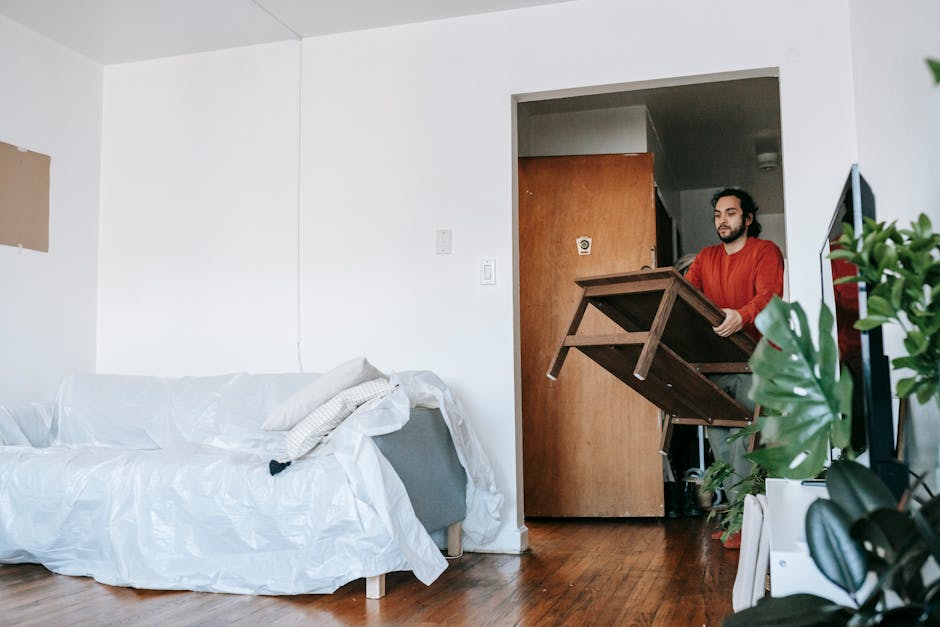 A man with dark hair and a beard, wearing a red long-sleeved shirt, is seen carrying a small wooden table through a doorway in a residential interior during a home relocation. The room has white walls, a light-colored sofa covered with a protective plastic sheet, and a wooden floor. In the foreground, a green houseplant is visible to the right, partially obstructing the view. Behind the man, the doorway leads to a corridor with a brown door and a light fixture on the ceiling. The scene captures the loading process as part of furniture transport in a house removal, with the man carefully handling the table to avoid damage. The natural lighting illuminates the space, and various packing materials or protective coverings may be seen on the furniture, reflective of the packing and moving procedures supported by Man with Van Upton, a professional removals service.