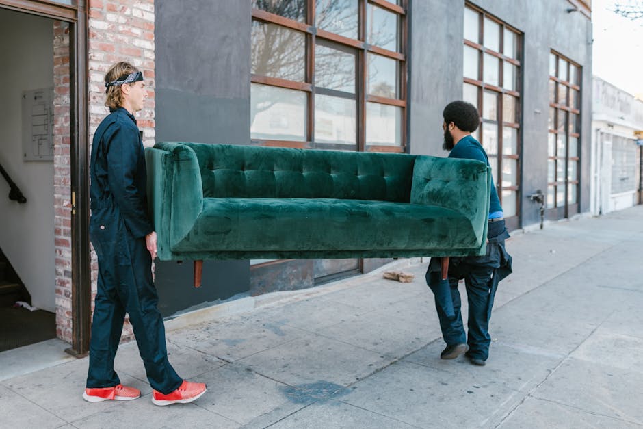 A man and woman involved in home relocation, positioned on a sidewalk outside a building with large windows and an industrial-style exterior. The woman, dressed in dark blue work overalls and red sneakers, stands facing the man, observing as he carefully carries a large, green upholstered sofa. The sofa, which appears to be in good condition with tufted detailing, is supported by wooden legs and is being moved from inside the property through an open doorway to the outside pavement. The man, wearing dark pants, a blue shirt, and black work shoes, grips the sofa firmly while walking along the sidewalk. The setting indicates a professional furniture transport process, with visible materials such as wrapping fabric or padding partially protecting the sofa. [COMPANY_NAME], Man with Van Upton, offers these relocation services, demonstrating careful handling during the loading and transport phase of a house move involving bulky furniture like sofas, which is a common aspect of professional removals and packing and moving tasks.