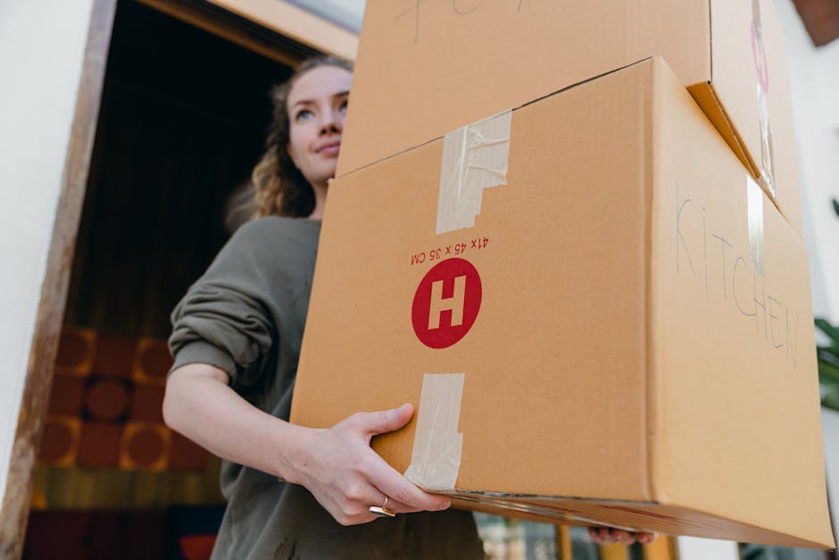 A woman with shoulder-length curly hair, wearing a grey long-sleeve top, is indoors holding a large cardboard box labeled 'KITCHEN' with masking tape. She stands near a doorway, preparing to load the box onto a moving vehicle or transport it within a house as part of a home relocation process. In the background, there is a dark interior with wooden paneling or furniture and some storage items. The box appears to be part of packing and moving activities coordinated by Man with Van Upton, a professional removals service specializing in house moves, wrapping furniture in cardboard and plastic for transport. The scene depicts her grip on the box with both hands, emphasizing careful handling during the loading or packing process of furniture and household items for a local move or house clearance.