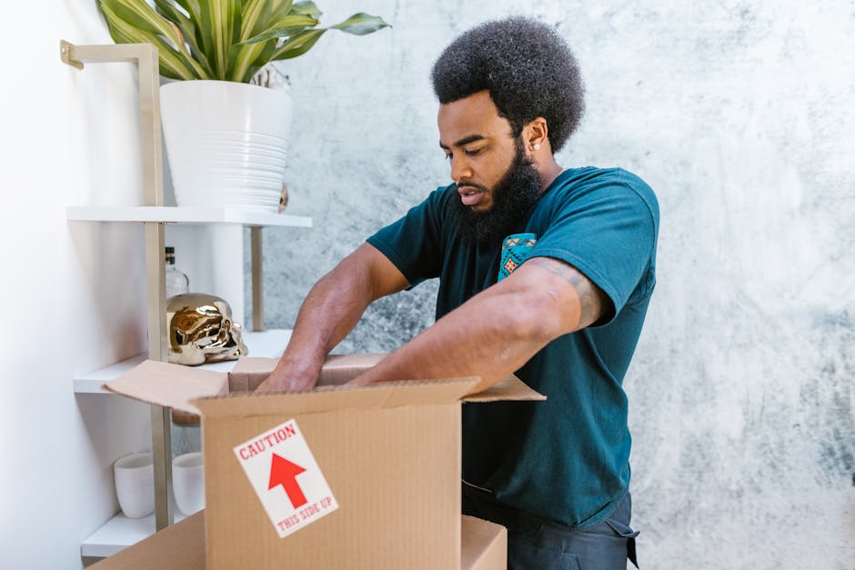 A woman with shoulder-length curly hair, wearing a grey long-sleeve top, is indoors holding a large cardboard box labeled 'KITCHEN' with masking tape. She stands near a doorway, preparing to load the box onto a moving vehicle or transport it within a house as part of a home relocation process. In the background, there is a dark interior with wooden paneling or furniture and some storage items. The box appears to be part of packing and moving activities coordinated by Man with Van Upton, a professional removals service specializing in house moves, wrapping furniture in cardboard and plastic for transport. The scene depicts her grip on the box with both hands, emphasizing careful handling during the loading or packing process of furniture and household items for a local move or house clearance.