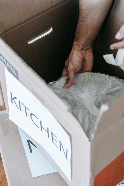 Inside a home, a person's hand is seen reaching into a cardboard box labeled 'KITCHEN' with a printed sign. The box contains packing materials such as bubble wrap, and it is situated on a surface near a brown door or cabinet. In the background, a vehicle, likely a van used for furniture transport and home relocation, is partially visible, indicating active loading or packing during a house move. The environment is well-lit, and additional packing materials such as plastic wraps and cardboard boxes are visible nearby. This scene represents the packing stage of a home removal, illustrating the careful handling and organization of belongings by Man with Van Upton as part of their moving services.
