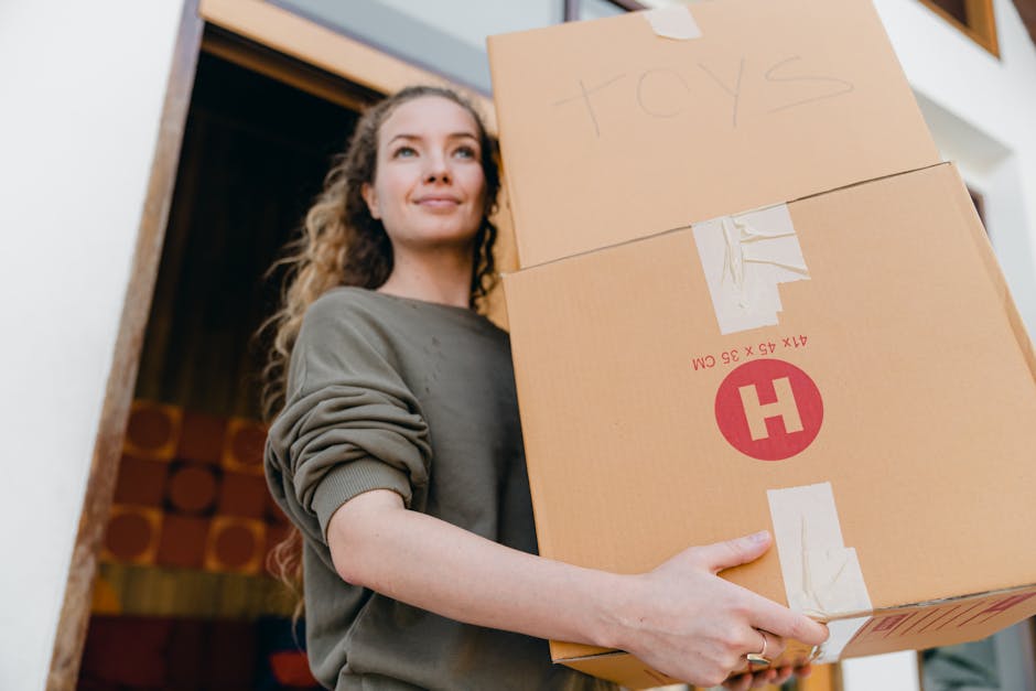 Inside a home, a person's hand is seen reaching into a cardboard box labeled 'KITCHEN' with a printed sign. The box contains packing materials such as bubble wrap, and it is situated on a surface near a brown door or cabinet. In the background, a vehicle, likely a van used for furniture transport and home relocation, is partially visible, indicating active loading or packing during a house move. The environment is well-lit, and additional packing materials such as plastic wraps and cardboard boxes are visible nearby. This scene represents the packing stage of a home removal, illustrating the careful handling and organization of belongings by Man with Van Upton as part of their moving services.