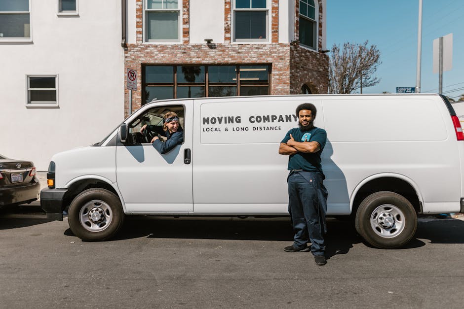 A man standing beside a white moving van with the label 'MOVING COMPANY LOCAL & LONG DISTANCE' on its side, parked on a city street in Upton. The van's sliding door is open, revealing a woman inside the vehicle who is holding a camera or device near a stack of cardboard boxes and wooden furniture wrapped in plastic and fabric blankets, ready for a home relocation. The street has other parked cars, and a backdrop of a brick and white facade building with multiple windows. The scene is captured during daylight, with clear weather and trees visible in the background. Manwithvanupton.co.uk provides professional removals services, assisting with packing and furniture transport for house moves, as part of efficient loading and moving logistics in Upton.