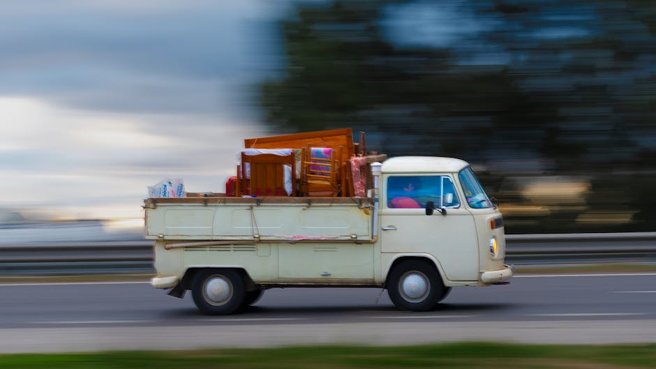 A cream-colored vintage van with a boxy shape is captured in motion along a road, with a blurred background indicating speed. The van's cargo area is loaded with various household items including wooden dining chairs, cardboard boxes wrapped in plastic film, and other packed belongings, suggesting a home relocation or moving process. Items are secured within the open back of the van, which is positioned on the right side of the image, with the road's surface and roadside fencing visible beneath. Inside the vehicle, visible elements include a red car seat and packaging materials, reinforcing the context of furniture transport and packing during a move. The scene is illuminated by natural daylight, with the blurred environment emphasizing the vehicle's movement. Man with Van Upton offers professional removals, supporting efficient and organized furniture transport as part of the home moving or packing and moving services.