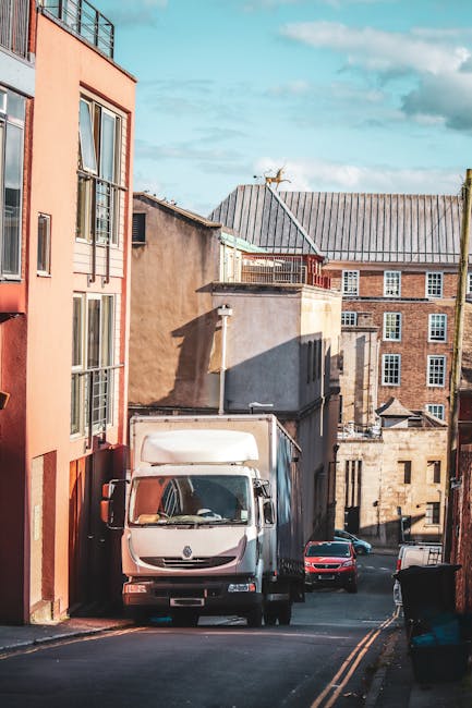 A white delivery van parked on a narrow urban street during daylight hours, with the rear doors closed and positioned close to the curb. The van is situated amidst a mixture of residential and commercial buildings featuring brick and stucco facades in various shades of beige, gray, and terracotta. In the background, additional vehicles including a red car are visible parked along the street which has a slight incline. The scene is lit by natural sunlight, casting shadows on the buildings and pavement. The street appears to be part of a home relocation process, with the van likely used by Man with Van Upton for furniture transport or packing and moving tasks, with nearby structures indicating potential access points for loading and unloading household items as part of house removals services.