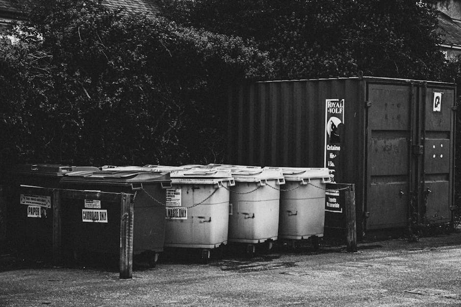 A row of waste and recycling containers situated outdoors on a paved surface, with the containers positioned in front of a large, dark shipping container. The waste bins include a reusable, marked 'E-WASTE ONLY' bin, a separate bin designated for paper recycling, and several large, white, plastic wheelie bins with closed lids, which are likely used for general waste disposal. The containers are arranged side by side, with some of the bins slightly overlapping or close together. There is a wooden post supporting a sign near the bins, and the area appears to be part of a residential or commercial premises, possibly involved in moving or disposal activities. The scene is captured in black and white, emphasizing the textures of the bins, the paved ground, and the background foliage, with natural daylight illuminating the scene. Man with Van Upton may occasionally handle waste management as part of their home relocation and furniture transport services, especially during packing or disposal phases.