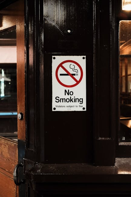 A close-up view of a black wooden pillar inside a property, featuring a white rectangular 'No Smoking' sign with a red circle and slash over a cigarette icon, and black text stating 'No Smoking' with smaller print 'Violators subject to fine.' The pillar is part of a doorway or interior wall, with a glimpse of a wooden counter or partition on the left side and warm ambient lighting reflecting off the surface. Visible in the background are wooden furniture and a window or glass panel, indicating a residential or commercial setting. The scene emphasizes the importance of restricted smoking areas in the context of house removals or moving processes, where safety and compliance are prioritised by companies such as Man with Van Upton, especially during packing, loading, and transportation of household items, furniture, and appliances.