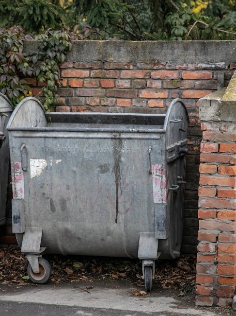 A row of waste and recycling containers situated outdoors on a paved surface, with the containers positioned in front of a large, dark shipping container. The waste bins include a reusable, marked 'E-WASTE ONLY' bin, a separate bin designated for paper recycling, and several large, white, plastic wheelie bins with closed lids, which are likely used for general waste disposal. The containers are arranged side by side, with some of the bins slightly overlapping or close together. There is a wooden post supporting a sign near the bins, and the area appears to be part of a residential or commercial premises, possibly involved in moving or disposal activities. The scene is captured in black and white, emphasizing the textures of the bins, the paved ground, and the background foliage, with natural daylight illuminating the scene. Man with Van Upton may occasionally handle waste management as part of their home relocation and furniture transport services, especially during packing or disposal phases.