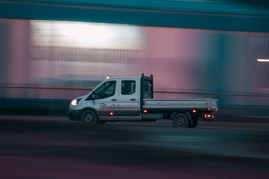 A silver flatbed van operated by Man with Van Upton is captured during the loading process on a street, with the vehicle in motion against a blurred urban background. The van's cargo area is visible, currently not carrying any items, and the vehicle's headlights are on. The scene suggests active packing and furniture transport related to home relocation or moving services. The pavement and surroundings are slightly lit by street lighting, and the vehicle is positioned parallel to the curb. This image reflects the logistics involved in house removals, highlighting the vehicle's role in swift and organized moving processes, ideal for urgent flat moves in Upton, as promoted on the relevant webpage. The background shows a structure with illuminated or reflective surfaces, emphasizing a city environment suitable for quick transportation services provided by Man with Van Upton.