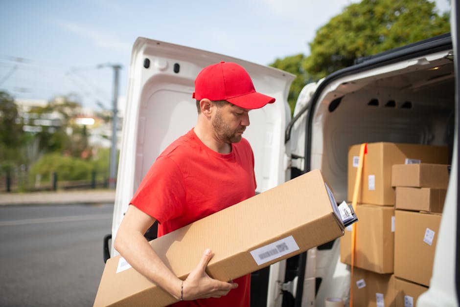 A man wearing a red cap and red t-shirt is seen loading a cardboard box into the back of a white moving van during daytime. The van's rear door is open, revealing multiple packed cardboard boxes inside, some secured with plastic wrap or packing tape. The man is standing outside on a paved road, with a suburban background including trees, houses, and utility poles visible in the distance. This scene captures the process of home relocation, involving furniture transport and packing during a moving service provided by Man with Van Upton, with the focus on careful handling and organization of items for efficient house removals and moving logistics.