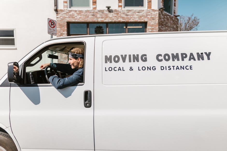 A silver flatbed van operated by Man with Van Upton is captured during the loading process on a street, with the vehicle in motion against a blurred urban background. The van's cargo area is visible, currently not carrying any items, and the vehicle's headlights are on. The scene suggests active packing and furniture transport related to home relocation or moving services. The pavement and surroundings are slightly lit by street lighting, and the vehicle is positioned parallel to the curb. This image reflects the logistics involved in house removals, highlighting the vehicle's role in swift and organized moving processes, ideal for urgent flat moves in Upton, as promoted on the relevant webpage. The background shows a structure with illuminated or reflective surfaces, emphasizing a city environment suitable for quick transportation services provided by Man with Van Upton.